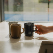 Three ceramic mugs in beige, brown, and black sit on a windowsill with a hand reaching toward the black mug. A bright window with blurred greenery and a pool is visible in the background.