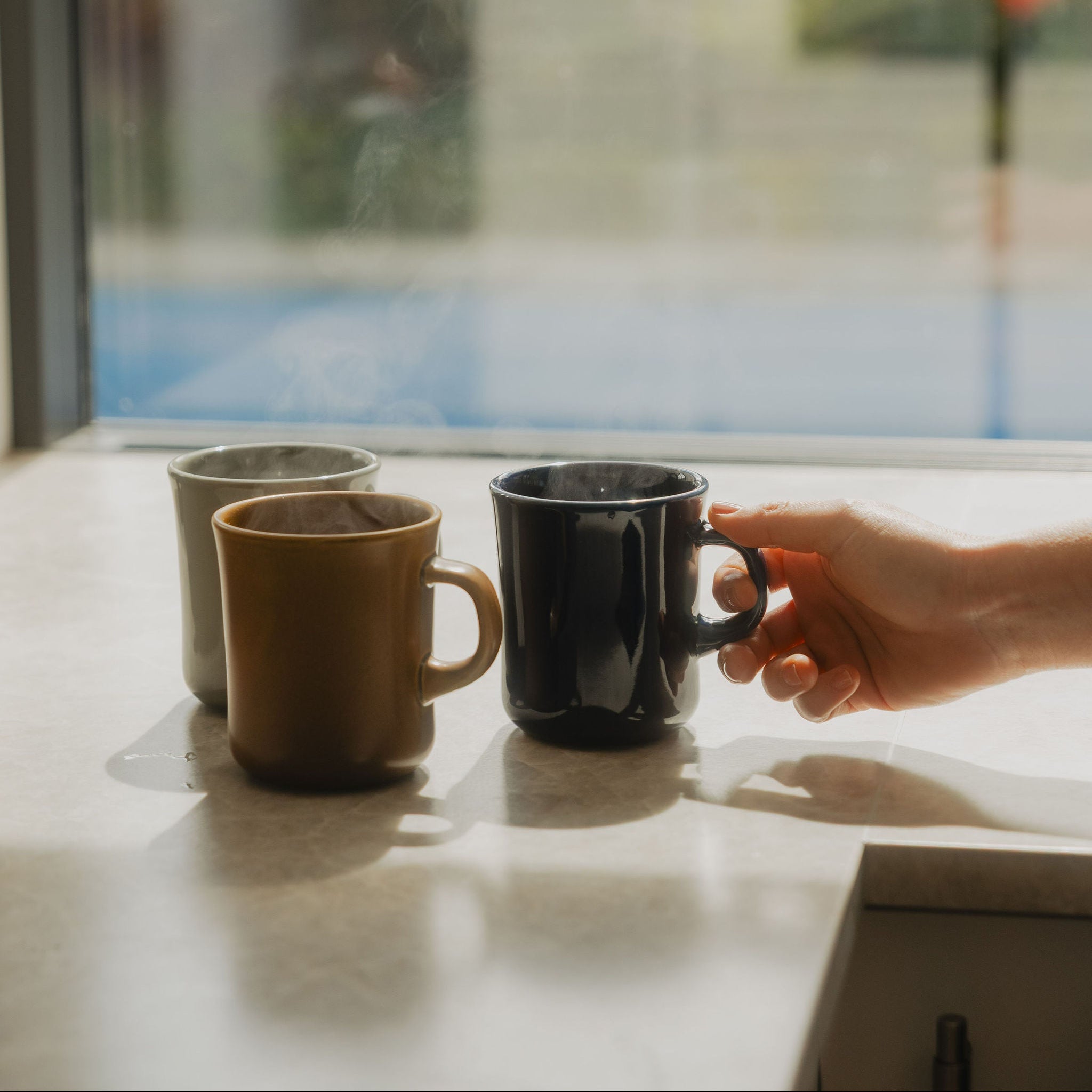 Three ceramic mugs in beige, brown, and black sit on a windowsill with a hand reaching toward the black mug. A bright window with blurred greenery and a pool is visible in the background.