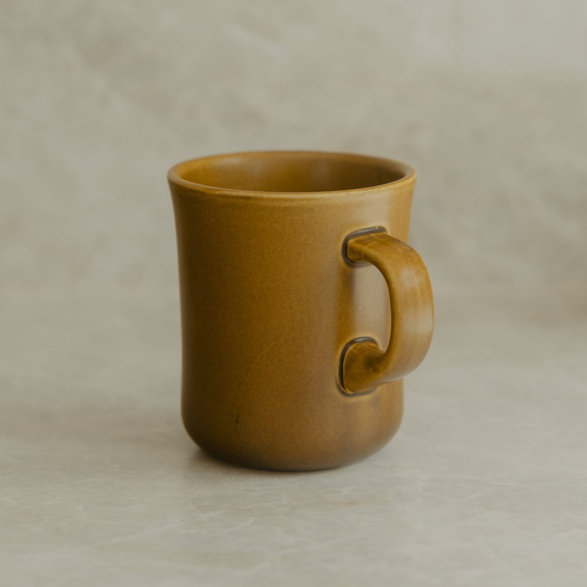 A single ceramic mug with a thick, rounded handle and a matte mustard-yellow finish, set against a neutral, textured beige background.
