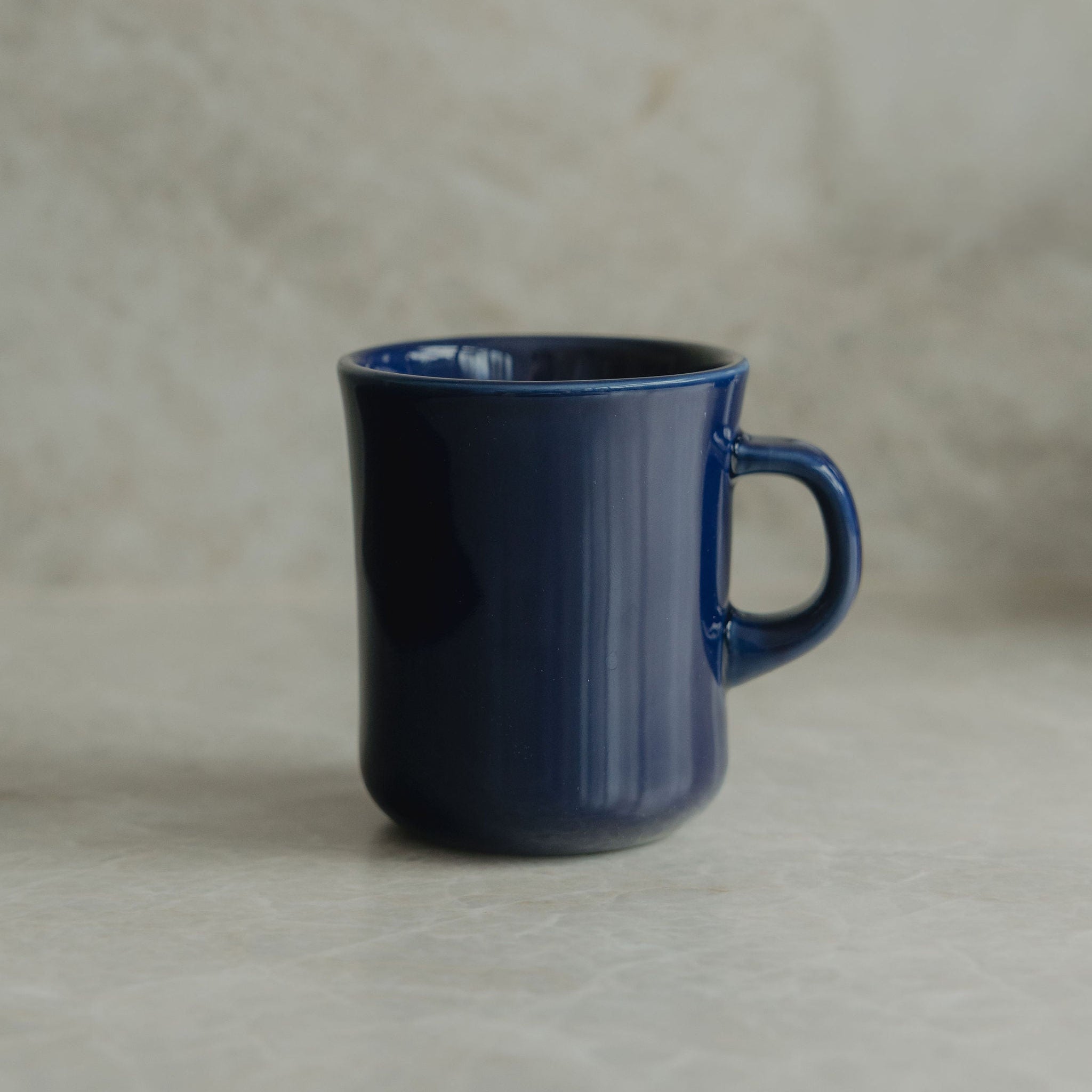 A dark blue ceramic mug with a matching handle, set against a neutral, textured beige background.