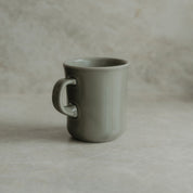 A matte sage green ceramic mug with a curved handle, standing on a textured beige stone surface against a matching stone background.