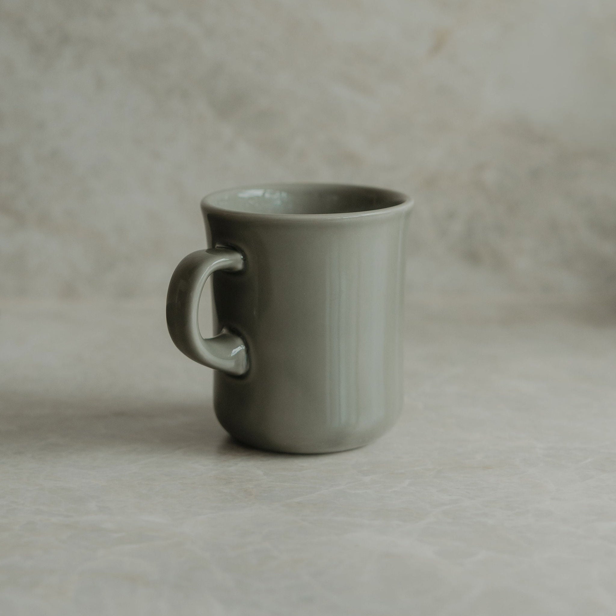 A matte sage green ceramic mug with a curved handle, standing on a textured beige stone surface against a matching stone background.