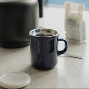 A dark navy blue ceramic mug with a stainless steel rim sits on a light stone countertop next to a black electric kettle and a white coffee bag, with a soft-focus window background.
