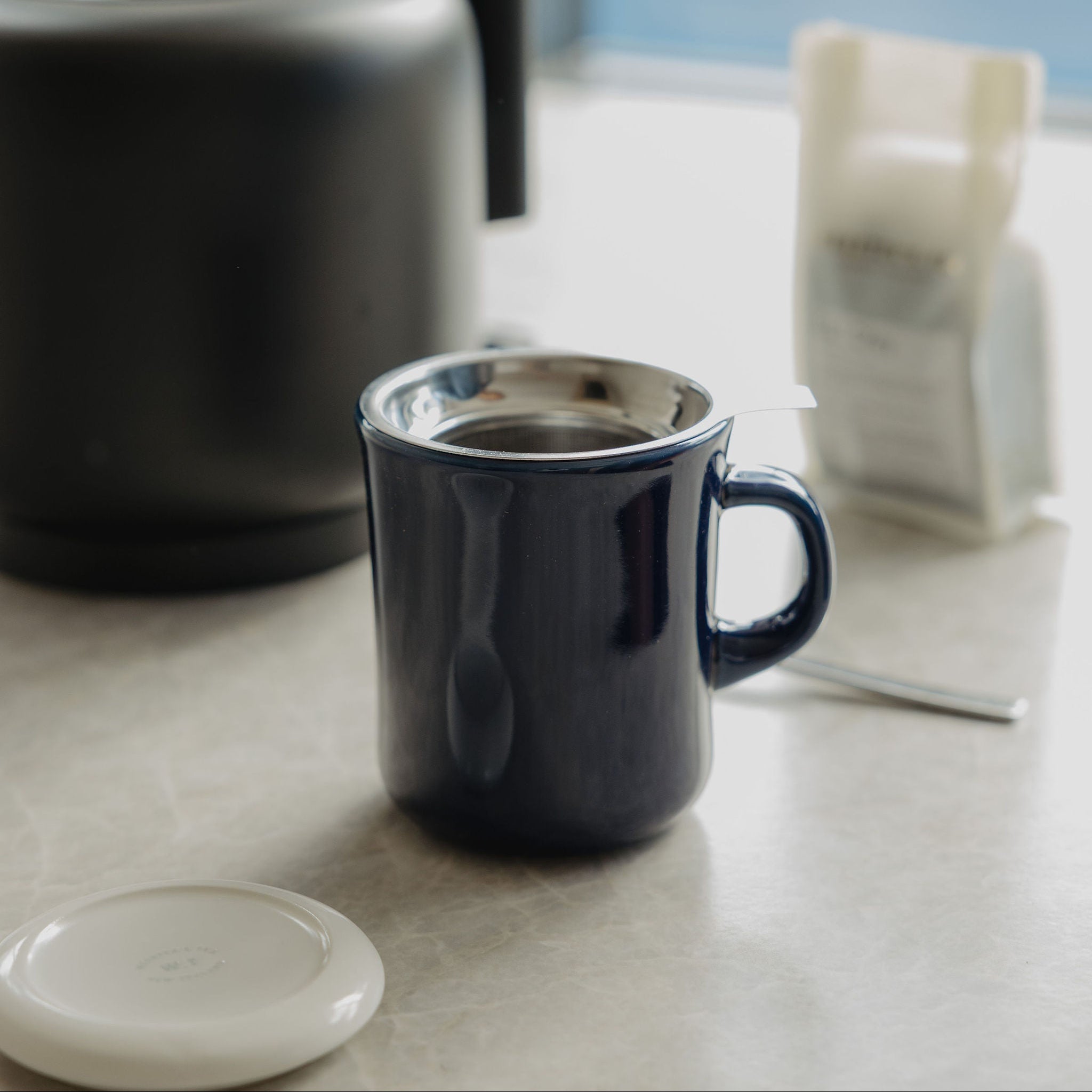 A dark navy blue ceramic mug with a stainless steel rim sits on a light stone countertop next to a black electric kettle and a white coffee bag, with a soft-focus window background.